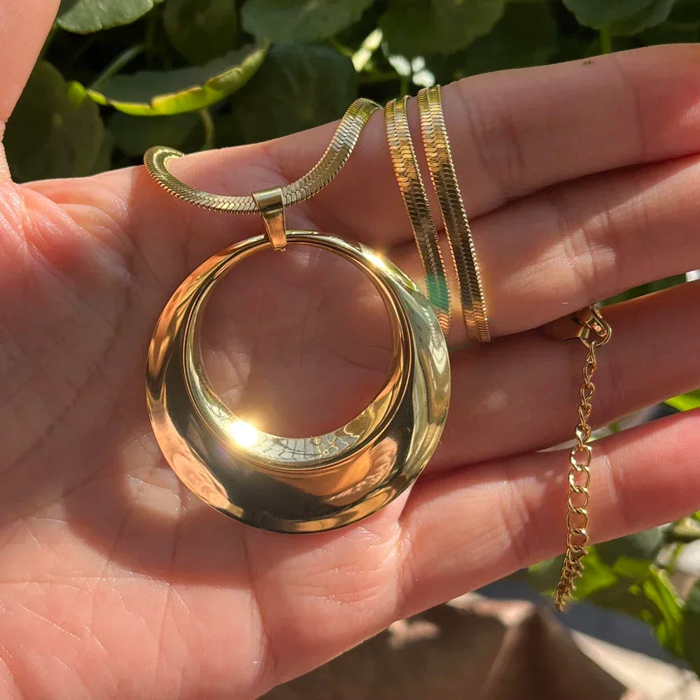 Gold necklace with a large circular pendant held in a hand against a natural background.